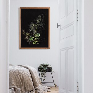 May include: A framed botanical print of a green plant with white flowers against a black background. The print is hanging on a white wall in a room with a wooden floor. A bed with a linen duvet is in the foreground. A small potted plant is on a black metal stand.