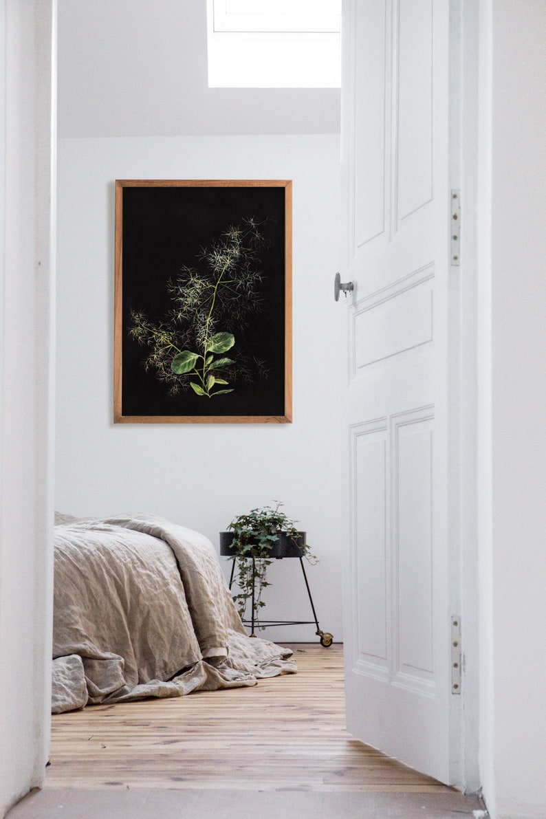May include: A framed botanical print of a green plant with white flowers against a black background. The print is hanging on a white wall in a room with a wooden floor. A bed with a linen duvet is in the foreground. A small potted plant is on a black metal stand.