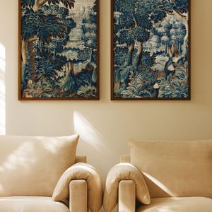 A pair of vintage verdure landscape prints in warm wood frames hangs above two cream armchairs in soft sunlight. The blue botanical tapestry design adds depth and timeless charm to the cozy living room wall art display.