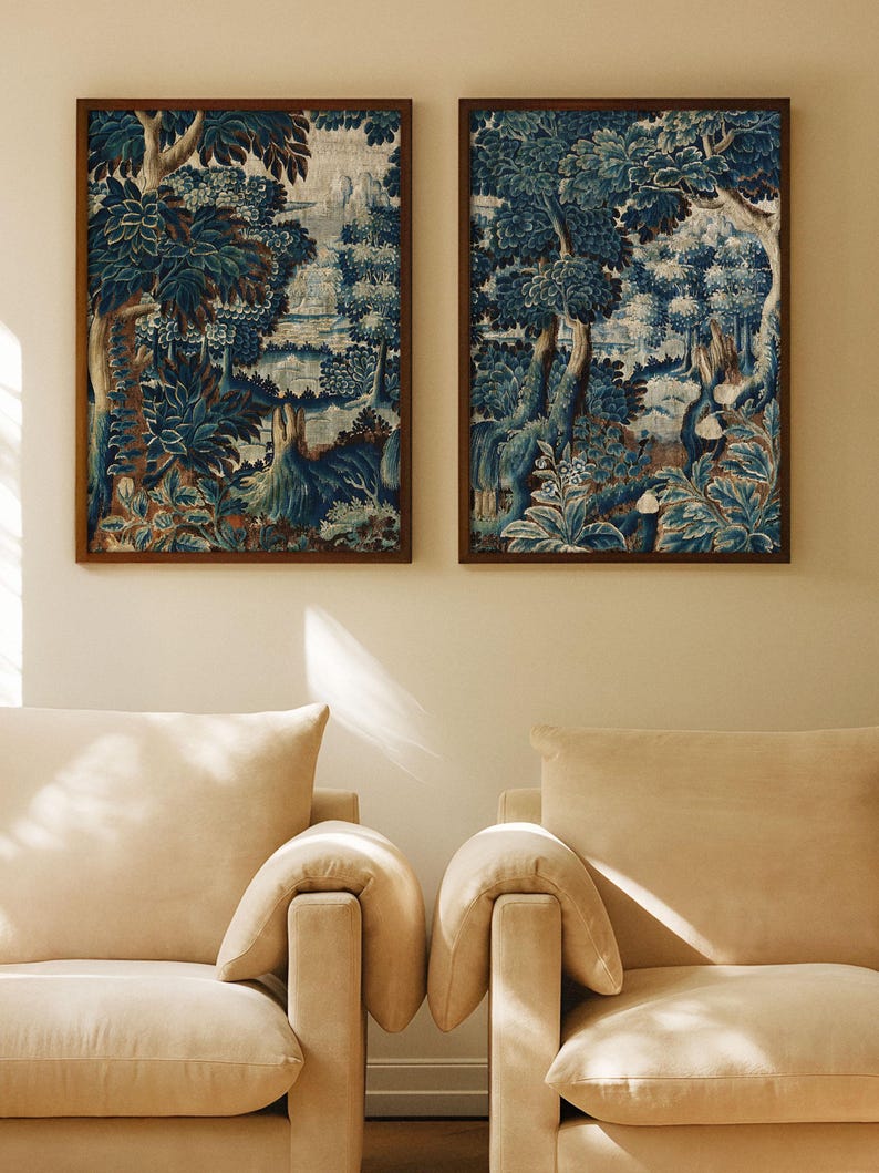 A pair of vintage verdure landscape prints in warm wood frames hangs above two cream armchairs in soft sunlight. The blue botanical tapestry design adds depth and timeless charm to the cozy living room wall art display.