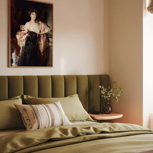 A bedroom with a soft green upholstered headboard, matching green bedding, and a decorative pillow. Above the bed hangs a framed portrait of a woman in a black and white gown with a red drape. A small bedside table holds a vase with greenery.