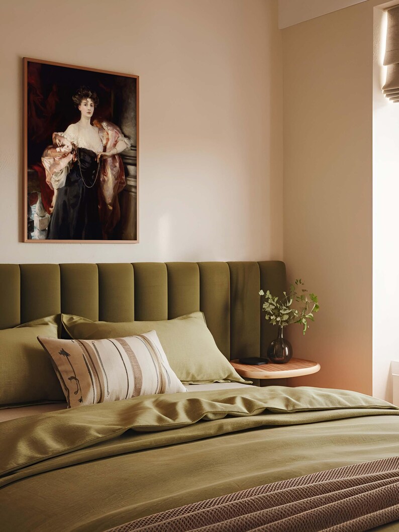 A bedroom with a soft green upholstered headboard, matching green bedding, and a decorative pillow. Above the bed hangs a framed portrait of a woman in a black and white gown with a red drape. A small bedside table holds a vase with greenery.