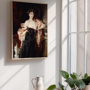 A framed portrait of a woman in a black dress with a pink shawl is hung on a wall next to a large window. Sunlight streams through the window, casting shadows. A small table with a white ceramic pitcher and a green potted plant are in the foreground.