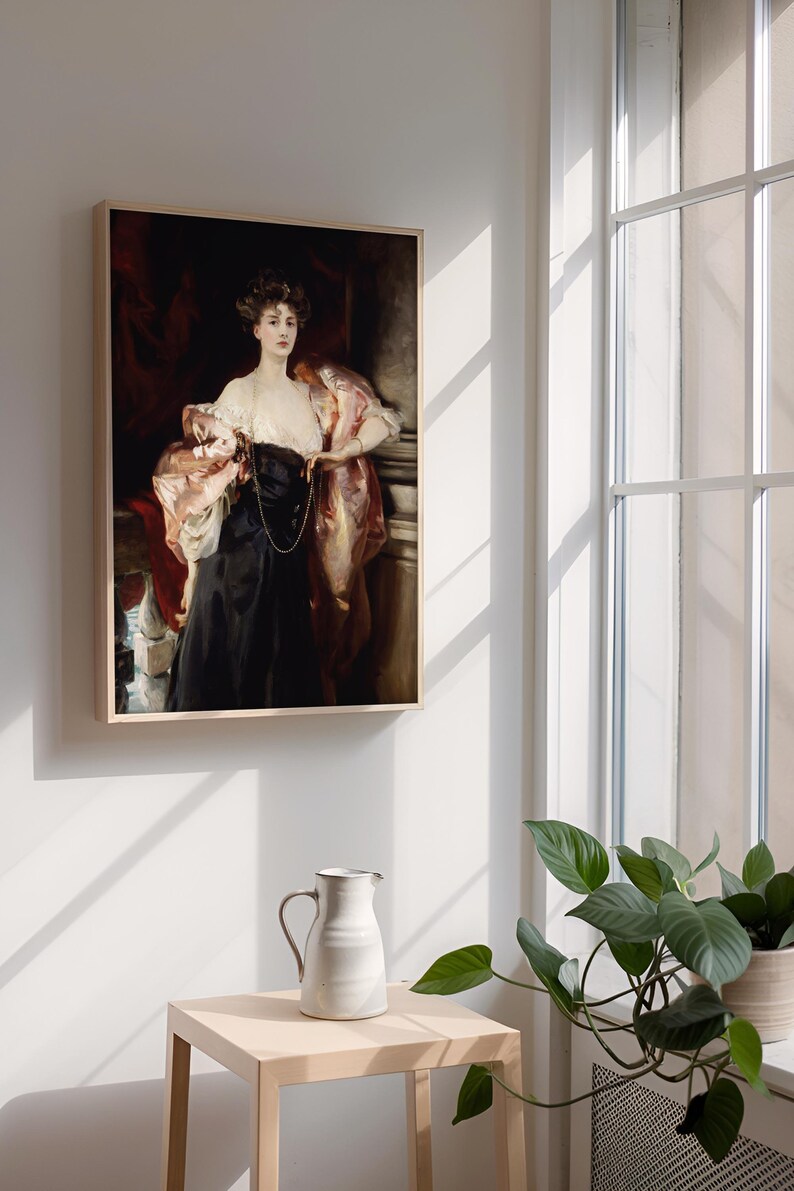 A framed portrait of a woman in a black dress with a pink shawl is hung on a wall next to a large window. Sunlight streams through the window, casting shadows. A small table with a white ceramic pitcher and a green potted plant are in the foreground.