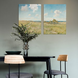 May include: Two framed landscape paintings of a grassy field with a dirt road leading to the ocean. The paintings are hanging on a light green wall above a black dining table with two gray chairs.
