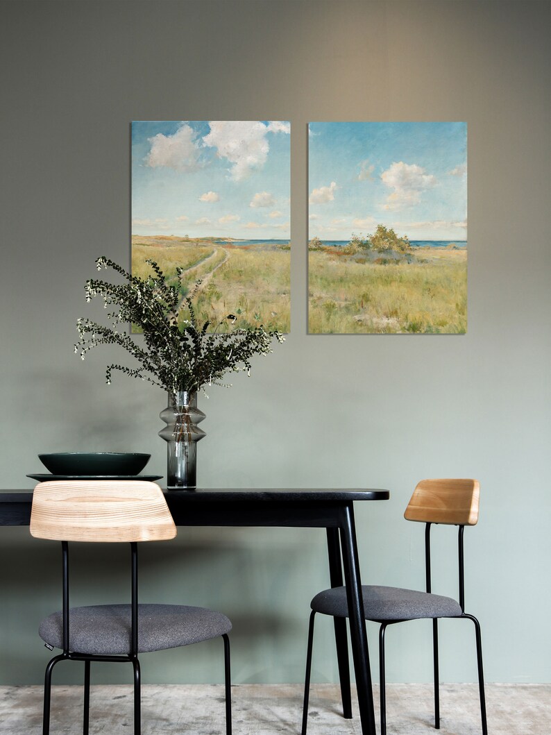 May include: Two framed landscape paintings of a grassy field with a dirt road leading to the ocean. The paintings are hanging on a light green wall above a black dining table with two gray chairs.