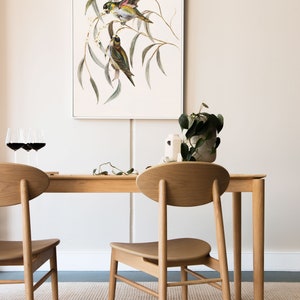 May include: Two wooden chairs with oval backs and a wooden table with a white framed print of two birds on a branch hanging above it.