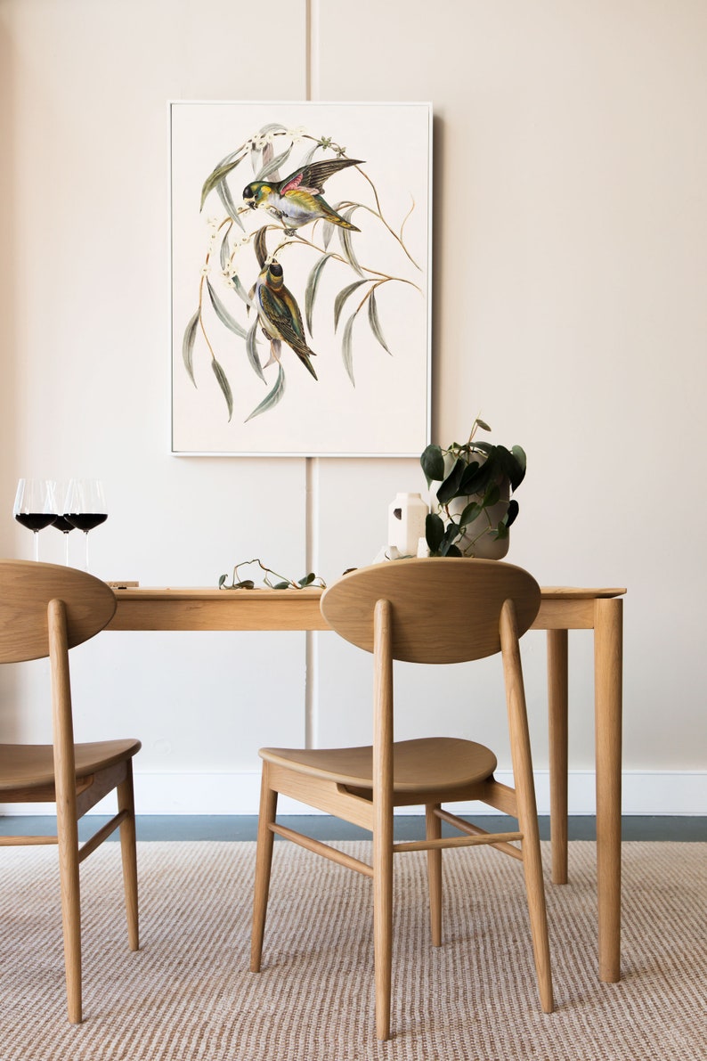 May include: Two wooden chairs with oval backs and a wooden table with a white framed print of two birds on a branch hanging above it.
