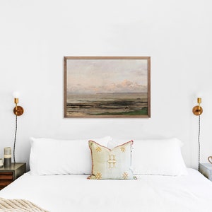 May include: A white bed with a white duvet and two pillows, one with a beige and gold patterned design. The bed is in a bedroom with white walls and a framed painting of a seascape above the headboard. There are two bedside tables with lamps on either side of the bed.