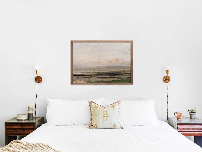 May include: A white bed with a white duvet and two pillows, one with a beige and gold patterned design. The bed is in a bedroom with white walls and a framed painting of a seascape above the headboard. There are two bedside tables with lamps on either side of the bed.