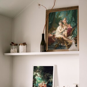 May include: Two framed paintings of classical art, one depicting a woman with two cherubs and the other a woman on a swing in a forest setting. The paintings are displayed on a white shelf with a glass bottle, two glass jars, and a wooden bowl.