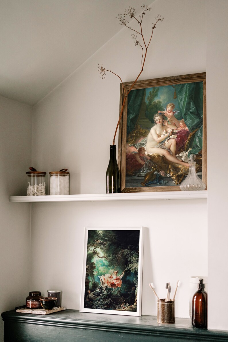 May include: Two framed paintings of classical art, one depicting a woman with two cherubs and the other a woman on a swing in a forest setting. The paintings are displayed on a white shelf with a glass bottle, two glass jars, and a wooden bowl.