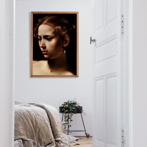 May include: A framed portrait of a woman with dark hair and a pearl earring. The woman is looking down and has a serious expression on her face. The portrait is hanging on a white wall in a room with a wooden floor.