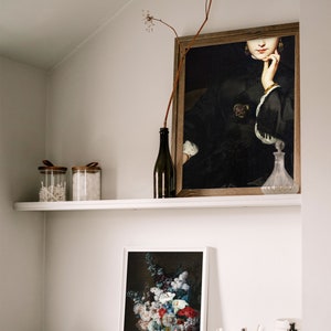 May include: A white shelf with a framed painting of a woman in a black dress, a framed print of a still life with flowers in a vase, two glass jars with lids, a bottle of wine, a glass bottle with a stopper, and two small bowls on a black shelf.