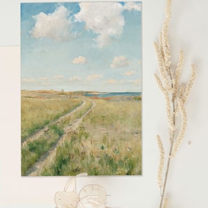 Art print lying flat on a white surface and surrounded by dried flowers. The print shows a landscape painting of a meadow with a path through it,  a seashore in the background and fluffy clouds in the blue sky.