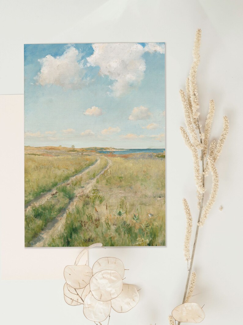 Art print lying flat on a white surface and surrounded by dried flowers. The print shows a landscape painting of a meadow with a path through it,  a seashore in the background and fluffy clouds in the blue sky.