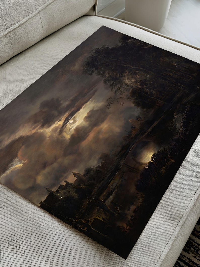 Unframed dark landscape art print lying flat on a beige sofa, showing moonlight breaking through dramatic clouds above a river and trees.
