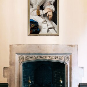 Antique fireplace above which hangs a golden frame showing detail of a victorian portrait of a woman wearing a white dress, golden rings and a golden bracelet with many gemstones.