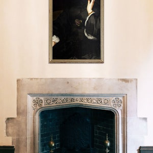 May include: A framed portrait of a woman in a black dress with a white collar, she is looking to the side and has her hand on her chin. The portrait is hanging on a white wall above a fireplace.