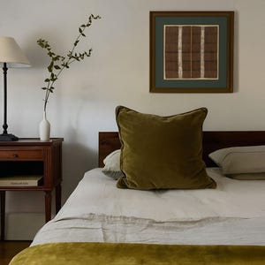 Framed vintage textile wall art with geometric stripes in earthy tones, hanging above a bed with olive green accents in a cozy bedroom.