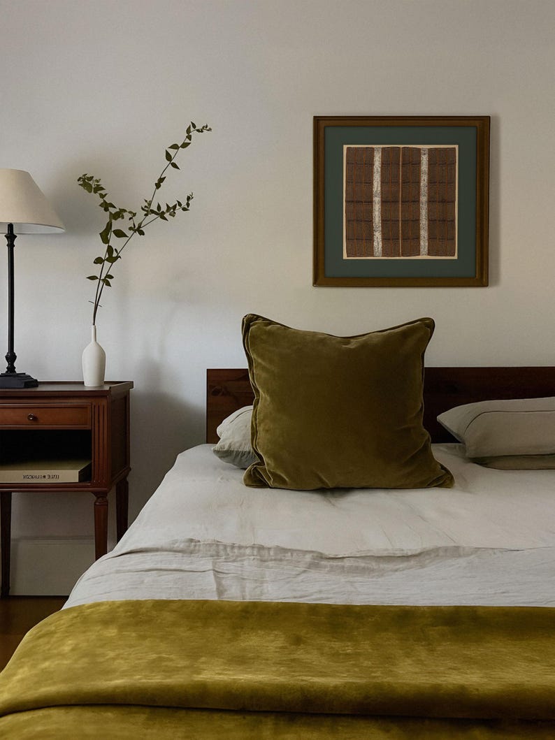Framed vintage textile wall art with geometric stripes in earthy tones, hanging above a bed with olive green accents in a cozy bedroom.