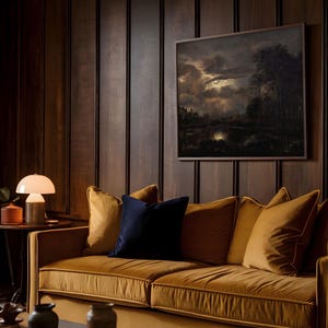 Framed dark moody landscape wall art with glowing moon over clouds, styled above a mustard velvet sofa with dark wood paneling and ambient lighting.