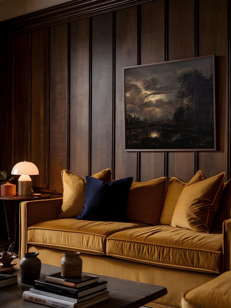 Framed dark moody landscape wall art with glowing moon over clouds, styled above a mustard velvet sofa with dark wood paneling and ambient lighting.