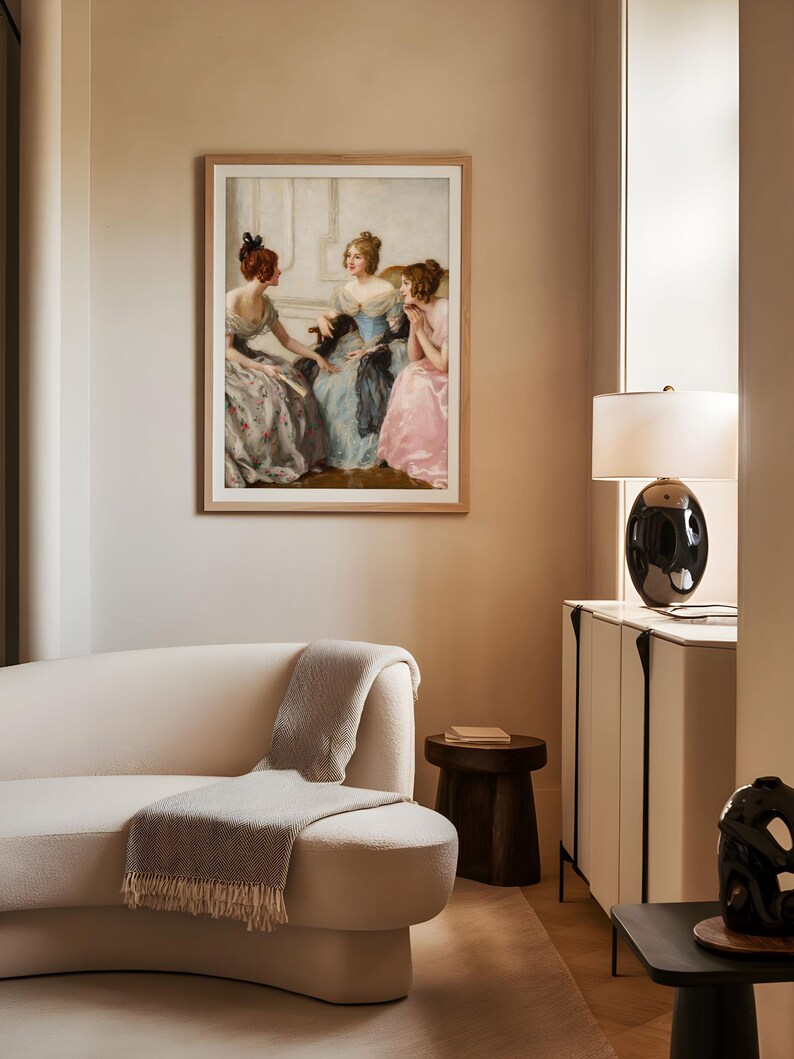 Framed art print of three Victorian women in pastel gowns conversing in an elegant interior, displayed above a curved cream sofa. The soft lighting and muted tones blend classical charm with contemporary living room decor.