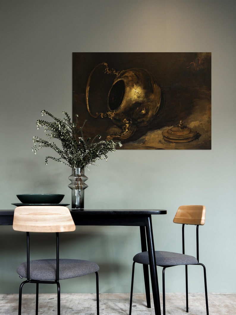 A vintage brass teapot art print is displayed on a sage green wall above a modern black dining table with wooden and gray chairs. A glass vase with greenery adds a fresh touch to the minimalistic, moody interior setting.