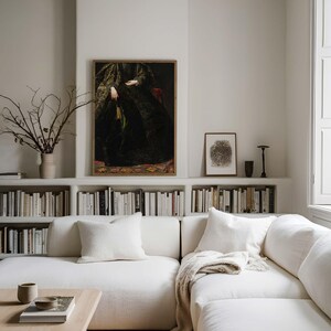 A framed vintage portrait of a woman in a dark, ornate gown is displayed on a white wall above a bookshelf in a modern living room. The room features a white sofa, neutral decor, and minimalistic accents, a wooden coffee table and soft throw blankets