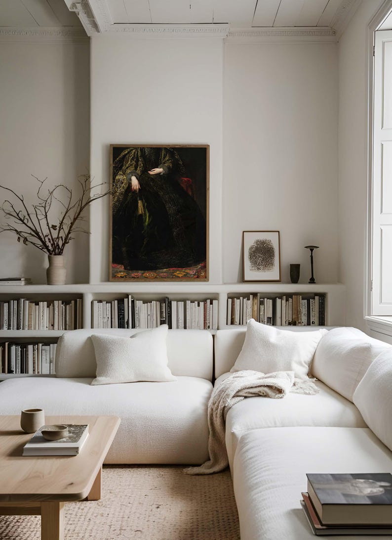 A framed vintage portrait of a woman in a dark, ornate gown is displayed on a white wall above a bookshelf in a modern living room. The room features a white sofa, neutral decor, and minimalistic accents, a wooden coffee table and soft throw blankets