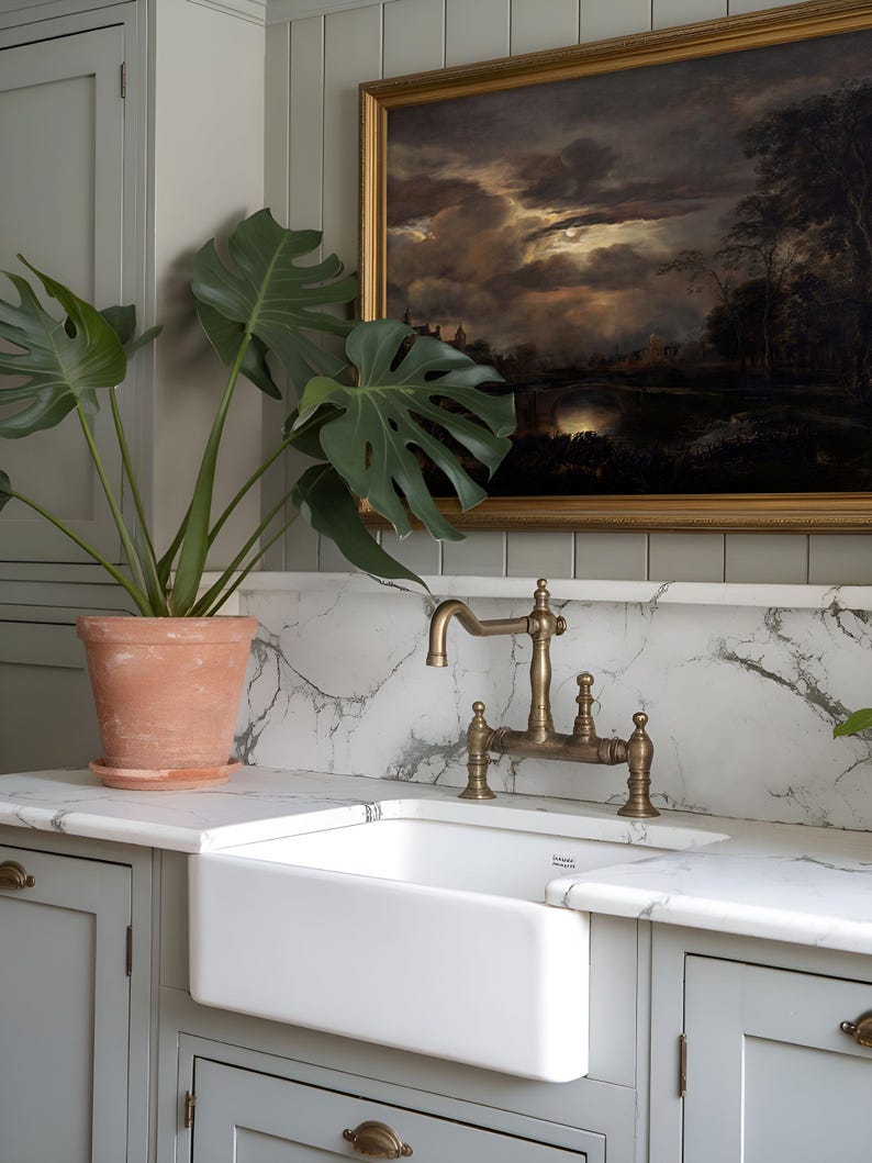 Dark landscape art print in a golden frame, displayed above a farmhouse sink with brass fixtures and a terracotta pot with green leaves in a kitchen setting.