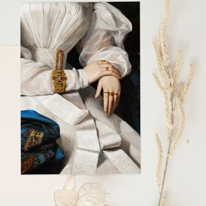 Art print laying flat on a white surface with dry flowers around it, the print shows a detail of a portrait painting of a woman wearing a white dress and many golden rings.
