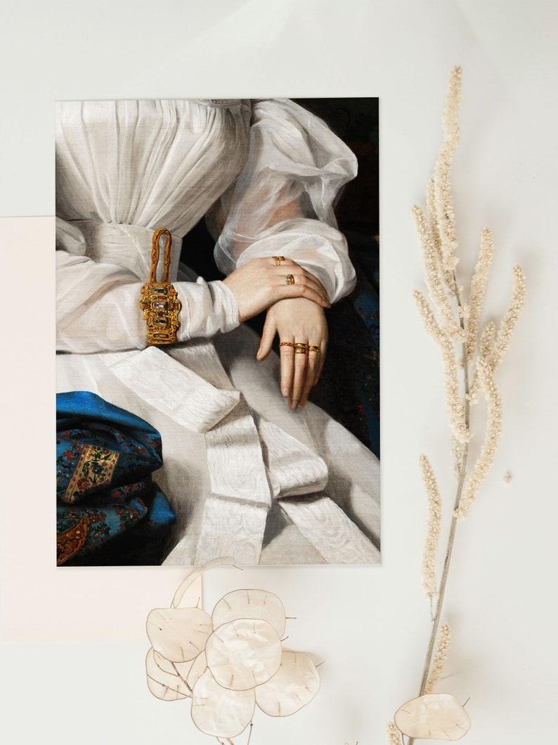 Art print laying flat on a white surface with dry flowers around it, the print shows a detail of a portrait painting of a woman wearing a white dress and many golden rings.