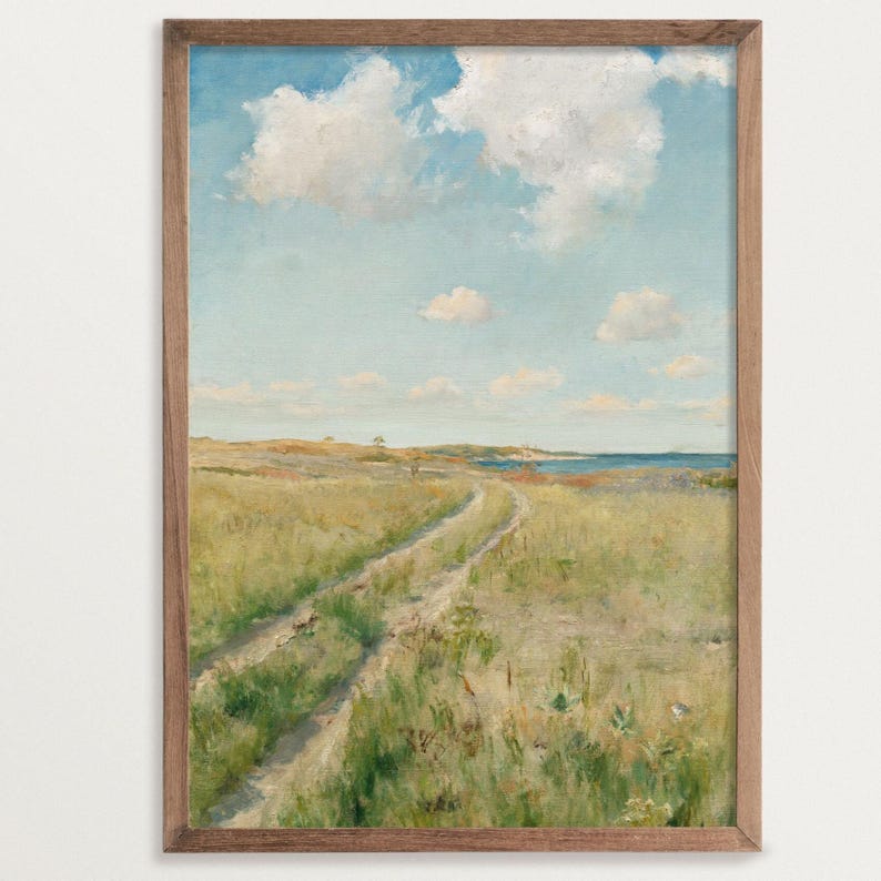 Landscape painting print in a wooden frame on a light grey wall. The print shows a landscape painting of a meadow with a path through it, a seashore in the background and fluffy clouds in the blue sky.