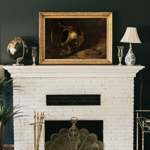 A dark, vintage-inspired art print of a brass teapot displayed above a white fireplace with a gold ornate frame. The dark green wall and antique decor, including a globe and candlesticks, create a timeless, moody atmosphere.