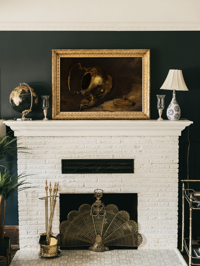 A dark, vintage-inspired art print of a brass teapot displayed above a white fireplace with a gold ornate frame. The dark green wall and antique decor, including a globe and candlesticks, create a timeless, moody atmosphere.