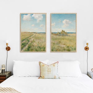 Bedroom with a large bed, beside it are two nightstands and lamps. On the wall above the bed are two art prints in frames showing a landscape painting of a meadow with a path through it, a seashore in the background and fluffy clouds in the blue sky.