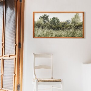 A cozy white room with a vintage panoramic landscape painting framed in warm wood above a rustic chair. The soft green meadow and gently faded brushwork evoke Wabi Sabi aesthetics, perfect for Japandi-inspired minimalist spaces with natural textures.