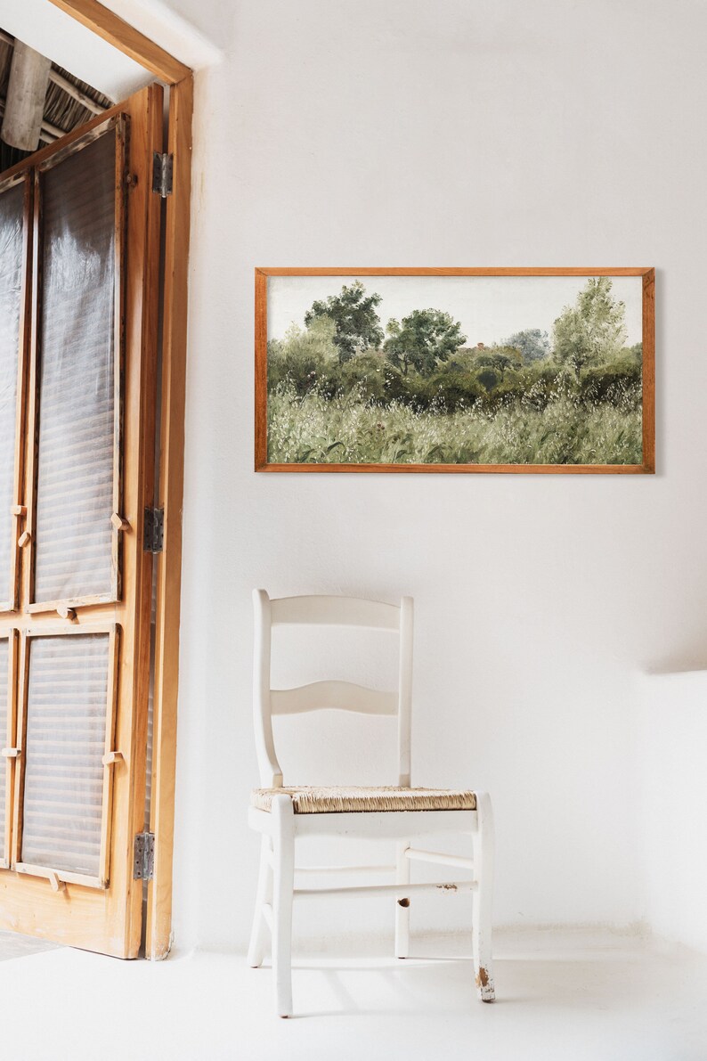A cozy white room with a vintage panoramic landscape painting framed in warm wood above a rustic chair. The soft green meadow and gently faded brushwork evoke Wabi Sabi aesthetics, perfect for Japandi-inspired minimalist spaces with natural textures.