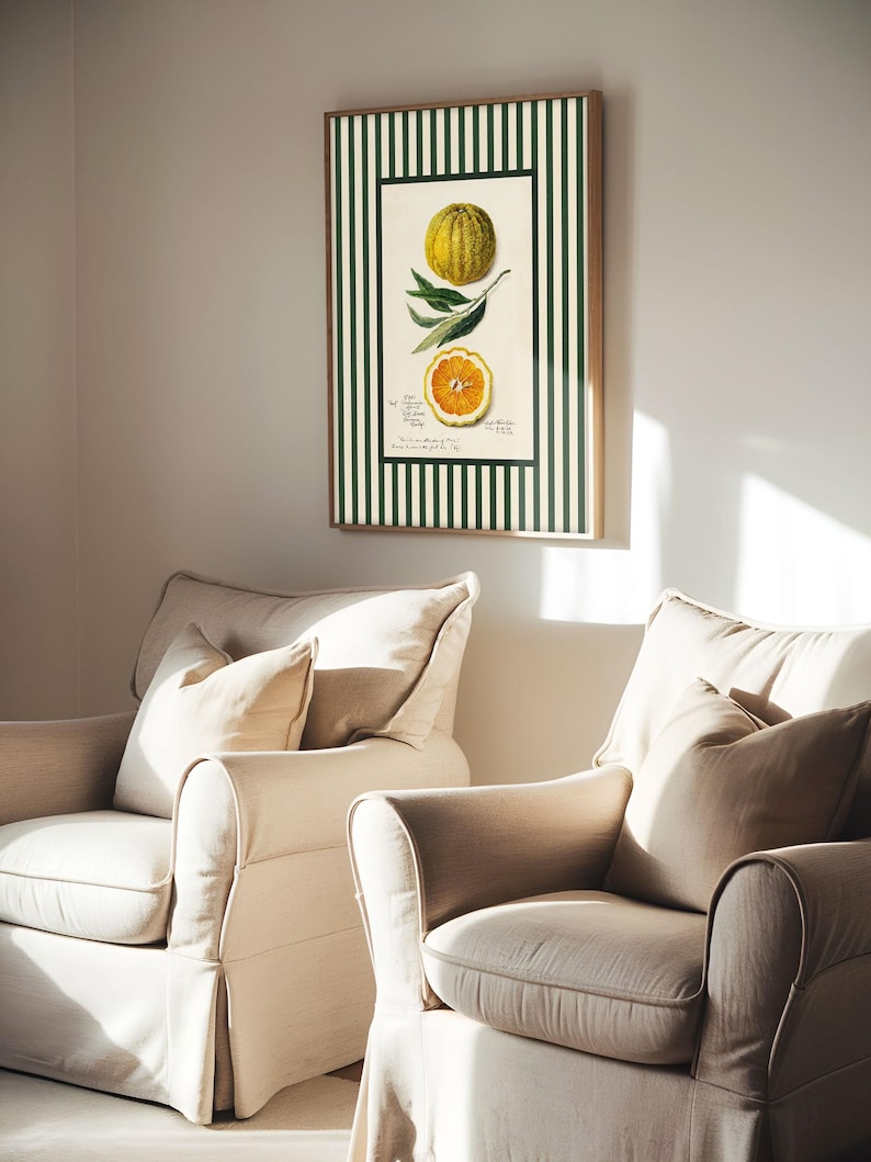 Green-striped orange print in a wooden frame hanging between two beige armchairs in a softly lit living room. A playful blend of traditional botanical and modern cottage aesthetic.