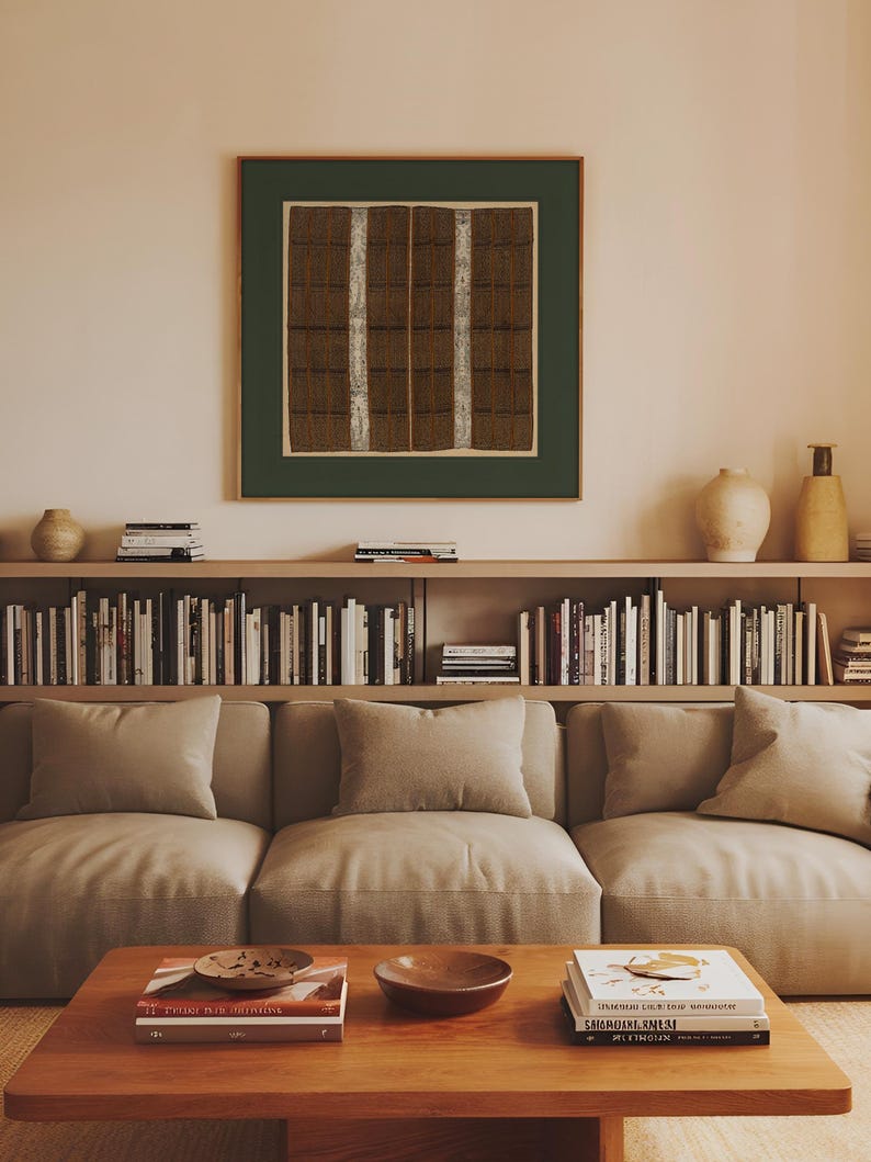 Vintage square textile wall art with intricate brown geometric patterns and cream vertical bands, surrounded by a dark green green border and hanging above a neutral sofa and built-in bookshelf in a warm modern living room.
