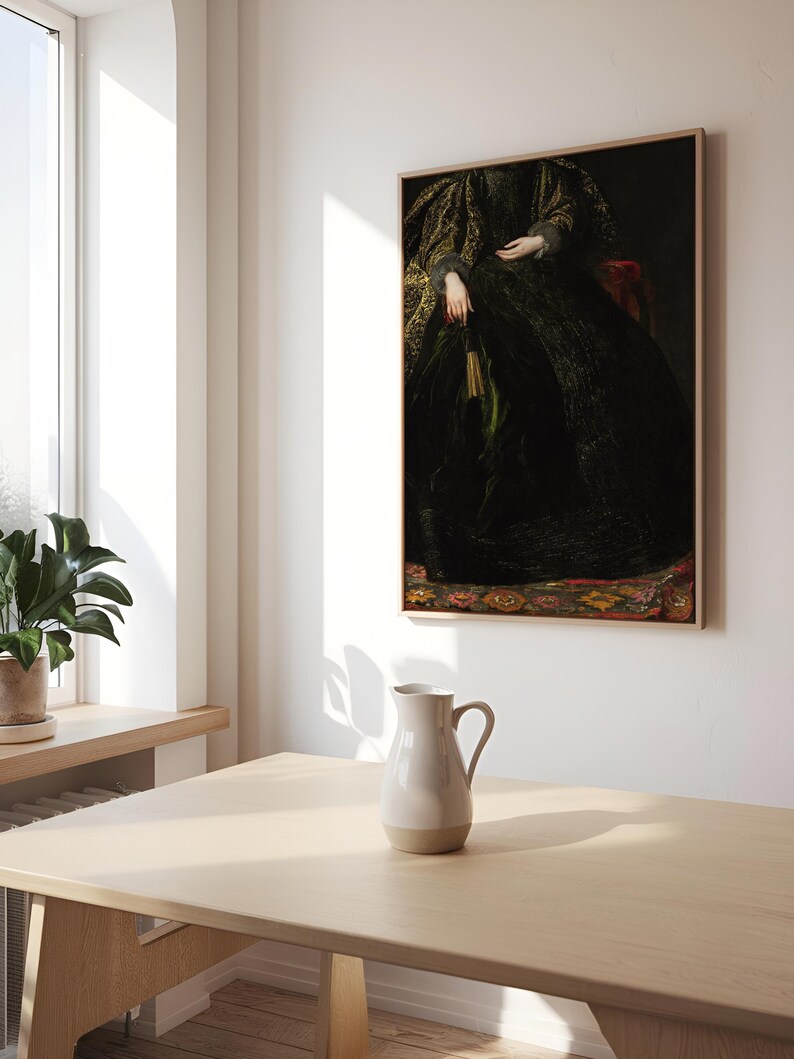 A framed portrait of a woman in a dark, intricate gown is hung on a white wall in a bright dining room. The wooden table below holds a simple ceramic pitcher, with sunlight streaming through the window, illuminating the minimalist modern decor.