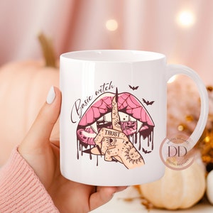 以下が含まれることがあります： 白いセラミック製のマグカップで、ピンクと黒の口のイラストが描かれており、牙とクモの巣のデザインが施されています。マグカップには「Basic Witch」という文字が筆記体で書かれています。手の指がリングで示されており、指には「Trust」という文字が書かれています。