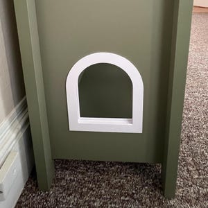 May include: A green wooden cat house with a white arched doorway. The cat house has a simple, modern design and is painted in a muted green color. The doorway is framed in white, creating a contrasting focal point.