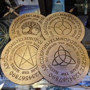 May include: Four circular wooden spirit boards featuring engraved alphabets, numbers 0-9, and the words YES, NO, MAYBE, and REPHRASE. Each board displays a unique central symbol: a tree, a four-pointed star, a pentagram, and a triquetra knot.