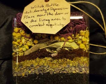 May include: A square glass bottle filled with layered botanical materials including black seeds, lavender, dried herbs, yellow chamomile flowers, and purple fibers. A tan paper tag is attached with twine, reading: Witch Bottle Anti Anxiety & Depression.