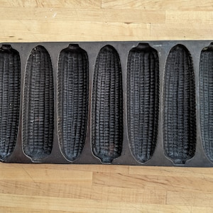 May include: A cast iron cornbread pan with six corn cob moulds. The pan is dark grey and sits on a light wood surface. The moulds are detailed to resemble corn cobs, perfect for baking. The pan has handles on each end.