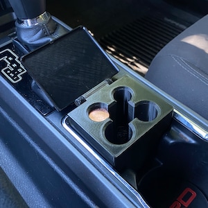 Puede incluir: Un portavasos de coche de plástico negro con una moneda en su interior. El portavasos está instalado en la consola central de un coche. La consola tiene una superficie de cuero negro y un ribete plateado. El portavasos tiene dos ranuras para vasos.