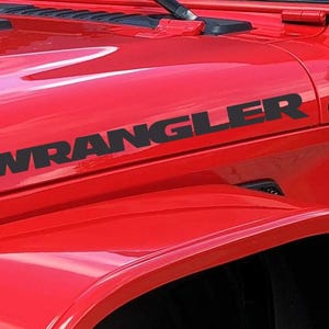 Puede incluir: Primer plano de un Jeep Wrangler rojo. La palabra "WRANGLER" se muestra en letras negras en el capó. La imagen destaca el acabado brillante y el diseño del vehículo.